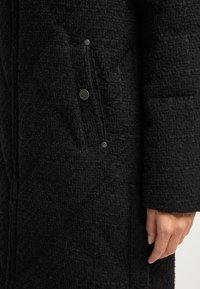 Sort quiltet frakke med tekstureret stof, en sidelomme med en sølvknap og et struktureret design med diagonale syninger.