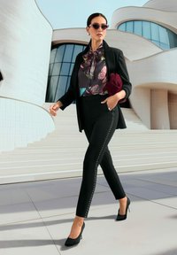 Zwarte gepersonaliseerde broek met studs aan de zijkanten, bloes met bloemenmotief en strik, en een getailleerde zwarte blazer. Accessoires zijn een bordeauxrode clutch en zonnebril.