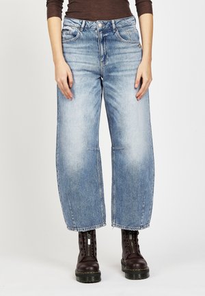 Frau trägt weit geschnittene, ausgewaschene blaue Jeans kombiniert mit dunkelbraunen Schnürstiefeln mit Reißverschluss vorne.