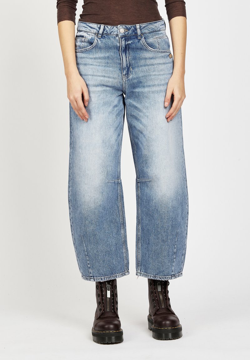 Frau trägt weit geschnittene, ausgewaschene blaue Jeans kombiniert mit dunkelbraunen Schnürstiefeln mit Reißverschluss vorne.