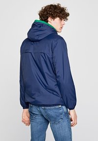 Giacca leggera blu navy con cappuccio e rivestimento verde. Polsini e orlo elastici. Presenta uno yoke posteriore e una texture liscia.