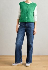 Gilet en maille verte porté sur une chemise blanche à manches longues, associé à un jean bleu à jambes larges et des chaussures blanches sans lacets. Fermeture simple à boutons sur le gilet.