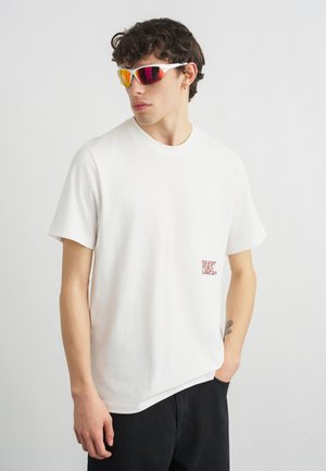 Fiatal férfi fehér pólóban, amelyen apró piros szöveg található, fekete nadrágban és tükröződő sport napszemüvegben, oldalra nézve, kezét a zsebében tartva.