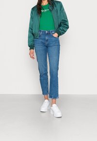 Grüne Bomberjacke mit gerippten Bündchen, kombiniert mit einem grünen T-Shirt mit weißem Text, blauen, taillierten Jeans und weißen Plateauschuhen.