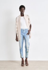 Beige offener Strickcardigan über einem weißen Oberteil, kombiniert mit hellblauen Jeans, die grüne Blumenstickereien an den Bündchen aufweisen, und braunen Absatzsandalen.