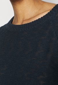 Tmavě modrý pletený svetr s kulatým výstřihem, s texturovaným povrchem a žebrovanými manžetami. Látka působí lehce a mírně průsvitně.