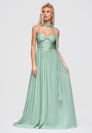 Femme blonde portant une robe longue sans manches vert clair avec une écharpe assortie drapée autour du cou.
