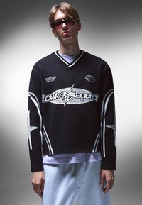 Svart, stickad tröja med vita detaljer, med V-ringning och stjärnmönster, ihop med gråa sweatpants. Bärs med tonade solglasögon.