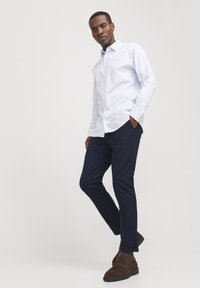 Weißes Hemd mit hellblauen Streifen, kombiniert mit navy-farbenen Slim-Fit-Hosen und braunen Wildleder-Schlüpfschuhen. Neutraler Hintergrund.