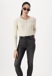 Kremfarget ribbet langermet cardigan med knapper foran, kombinert med høy midje svarte skinny jeans. Modell bruker svarte solbriller.