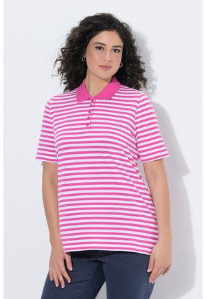 Femme aux cheveux bouclés foncés portant un polo rayé rose et blanc à manches courtes et un pantalon foncé, debout devant un fond blanc.