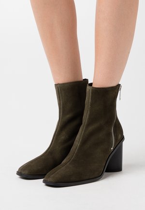 Olivgrüne Wildleder-Stiefeletten mit quadratischer Zehenform, seitlichem Reißverschluss und massiver schwarzer Absatz. Glatte Textur mit minimaler Verzierung.