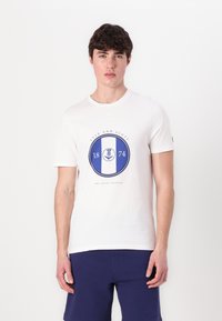 Balta trumpa rankovėmis marškiniai su mėlynu apvaliu logotipu, kuriame užrašyta "LYLE AND SCOTT," "1874" ir šeivamedžio dizainas centre. Lygus paviršius.