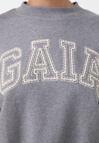 Grijze sweatshirt met verhoogde, geborduurde witte letters die "GAIA" spellen. Heeft een geribbelde kraag en een zachte, gestructureerde stof.
