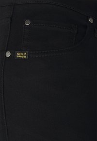Jeans de mezclilla negra que presentan una vista en primer plano de un bolsillo frontal, herrajes plateados y una pequeña etiqueta amarilla y negra que dice "tiger of Sweden."