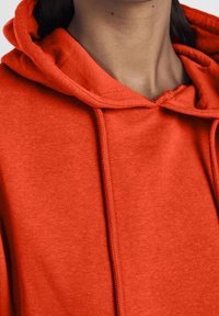 Röd hoodie med dragsko, mjukt material och avslappnad passform, med synliga sömmar längs sömmarna.