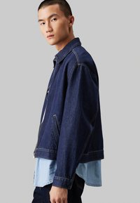 Giacca di jeans blu scuro con un taglio ampio, dotata di chiusura con zip, tasche laterali e cuciture a contrasto. Indossata sopra una camicia azzurra chiara.