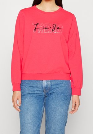 Kobieta w jasno-różowej bluzie z długim rękawem z napisami „Trend” i „Live joy”, w połączeniu z niebieskimi dżinsami, widziana od szyi do bioder.