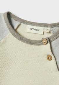 Camiseta en color crema claro y gris que presenta una tela texturizada, mangas raglán y acentos de botón de madera cerca del cuello, con etiqueta de marca visible.
