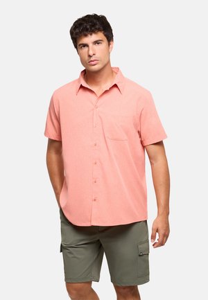Joven con una camisa salmonada de manga corta con botones y pantalones cortos cargo verde oliva, de pie frente a un fondo blanco liso.