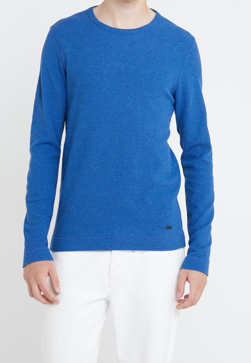 Blå långärmad t-shirt i ett strukturerat stickat tyg. Har en rund halsringning och en liten logotags vid fållen, matchad med vita byxor.