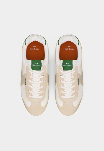 Paire de baskets beige et blanche avec une étiquette verte sur la languette et des lacets blancs, comportant la marque "PS Paul Smith" à l'intérieur et sur la languette.