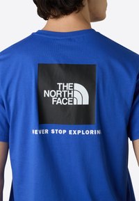 Blå kortærmet t-shirt med et sort firkantet grafisk design og hvid tekst samt The North Face-logoet på ryggen. Glat bomuldsstof.