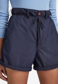 Námořnicky modré šortky vyrobené z hladkého materiálu s tkaničkou a zapínáním na knoflík v pase; mají ohrnuté manžety pro ležérní vzhled.