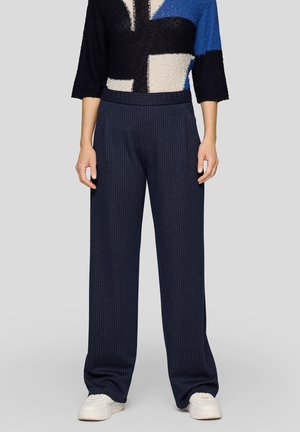 Person trägt marineblaue, breit geschnittene Anzughose mit Nadelstreifen, einen Pullover mit Farbblöcken und weiße Turnschuhe und steht vor einem einfarbigen grauen Hintergrund.