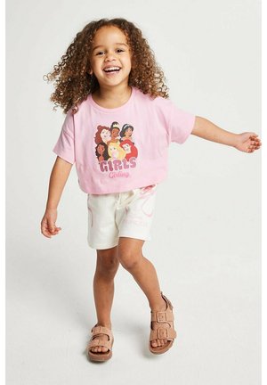 Lächendes Mädchen mit lockigem Haar trägt ein rosa Disney-Prinzessinnen-Crop-Top, weiße Shorts mit pinken Mustern und braune Sandalen und steht mit ausgestreckten Armen.