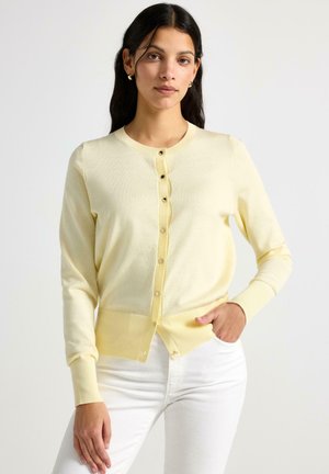 Kvinde med langt mørkt hår iført en lysegul knapcardigan og hvide bukser, stående med den ene hånd i lommen mod en hvid baggrund.