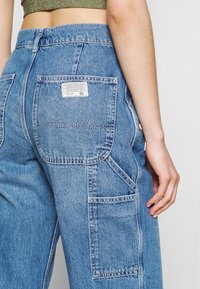 Ljust blå denim cargo byxor med stora fickor och en etikett på baksidan. Har en lös passform och strukturerad textil.