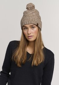 Gehaakte beanie in een gestructureerde bruine tint met een pompon bovenop en een leren label; gedragen met een donkere trui. Zacht en comfortabel materiaal.