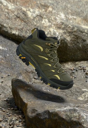 Zielony i czarny but trekkingowy Merrell z podeszwą Vibram zawieszony nad skalistym terenem.