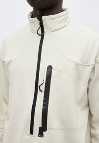 Κοντινό πλάνο ατόμου που φοράει μπεζ μπουφάν με ψηλό γιακά, μαύρο κάθετο φερμουάρ και μπροστινή τσέπη στο στήθος με μαύρο αδιάβροχο φερμουάρ.