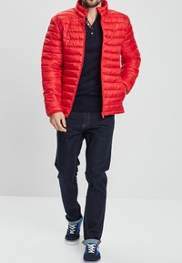 Chaqueta acolchada roja con secciones acolchadas horizontales, cuello alto, cierre de cremallera, combinada con una camisa azul marino y jeans oscuros, usada con zapatillas azules.