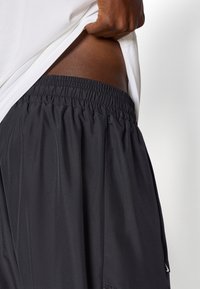 Calções atléticos pretos com uma cintura elástica e uma textura suave, apresentando um ajuste solto. Uma camisa branca cobre parcialmente a cintura.