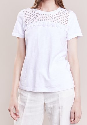 Biały krótki top z koronką szydełkową i frędzlami na piersi, noszony z jasnymi, teksturowanymi spodniami.