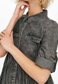 Tummanharmaa farkkumekko, jossa on napillinen etuosa, lyhyet kääritut hihat, kaksi rintataskua ja kerrostettu vyötärö rennon istuvuuden takaamiseksi.