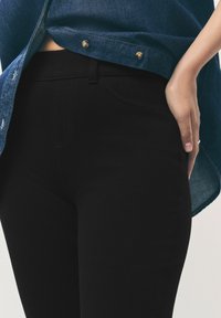 Schwarze Jeans aus Denim mit schmalem Schnitt, glatter Textur und klassischem Fünf-Taschen-Design, kombiniert mit einem blauen Denim-Hemd in der Taille.
