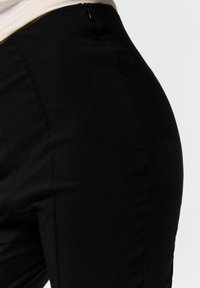 Gros plan d'une personne portant un pantalon noir taille haute avec des coutures visibles et une fermeture éclair latérale sur un fond clair uni.