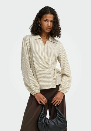 Frau mit lockigem Haar trägt eine beige gestreifte Wickelbluse und braune Hose und hält eine schwarze Lederhandtasche vor einem einfarbigen Hintergrund.