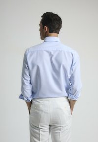 Camisa de manga larga azul claro con cuello abotonado, mangas enrolladas y textura suave, llevada con pantalones blancos.
