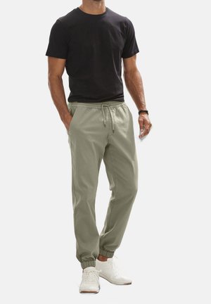Man draagt een zwart T-shirt met korte mouwen, licht olijfgroene joggingbroek met trekkoord, witte sneakers en een zwarte polsband, staat met handen in de zakken.