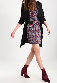 Zwarte omslagjas over een jurk met een bloemmotief in blauw, rood en wit, gecombineerd met mahoniekleurige enkellaarsjes. De outfit heeft een gelaagde uitstraling.