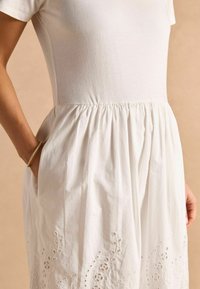 Weißes Baumwollkleid mit kurzen Ärmeln, das eine geraffte Taille und ein besticktes Blumendesign am Saum aufweist. Seitentaschen sind sichtbar.