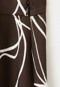 Tissu marron avec des lignes courbes blanches abstraites et un tirette de fermeture éclair noire au centre sur un matériau texturé.