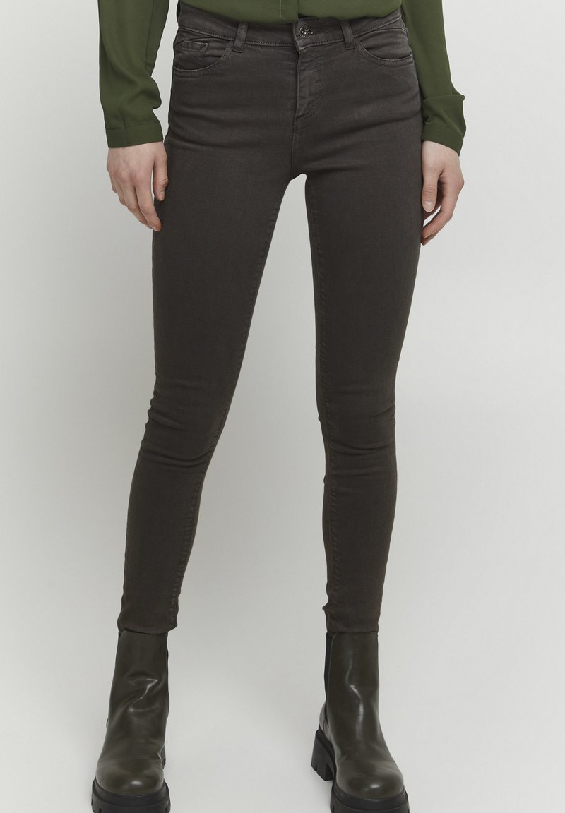 Jeans skinny marron en denim, dotés d'un design classique à cinq poches, avec des coutures discrètes, associés à des bottes vertes jusqu'aux genoux.