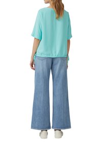 Helles blaues, kurzärmliges Blusenshirt mit gerafftem Bund und Bindedetails, kombiniert mit weiten, hellen Jeans und weißen Sneakern.
