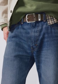 Jean en denim bleu foncé avec surpiqûres orange, assorti à une ceinture en cuir marron avec une boucle argentée et une chemise à carreaux en dessous.